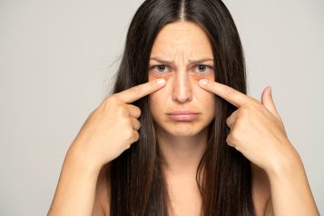 A,Young,Unhappy,Woman,Touches,Her,Undereyes,On,A,Gray