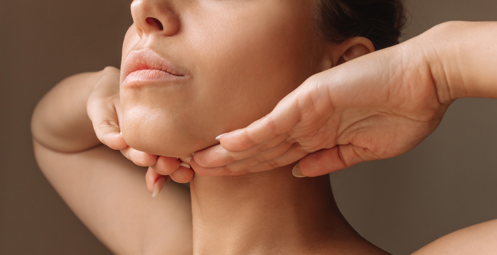 cropped shot of young caucasian woman touching under the chin with hands massaging her face on dark brown background