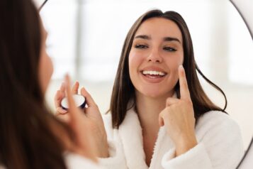 Cheerful,Young,Woman,Wearing,Bathrobe,Applying,Moisturizer,Cream,On,Her