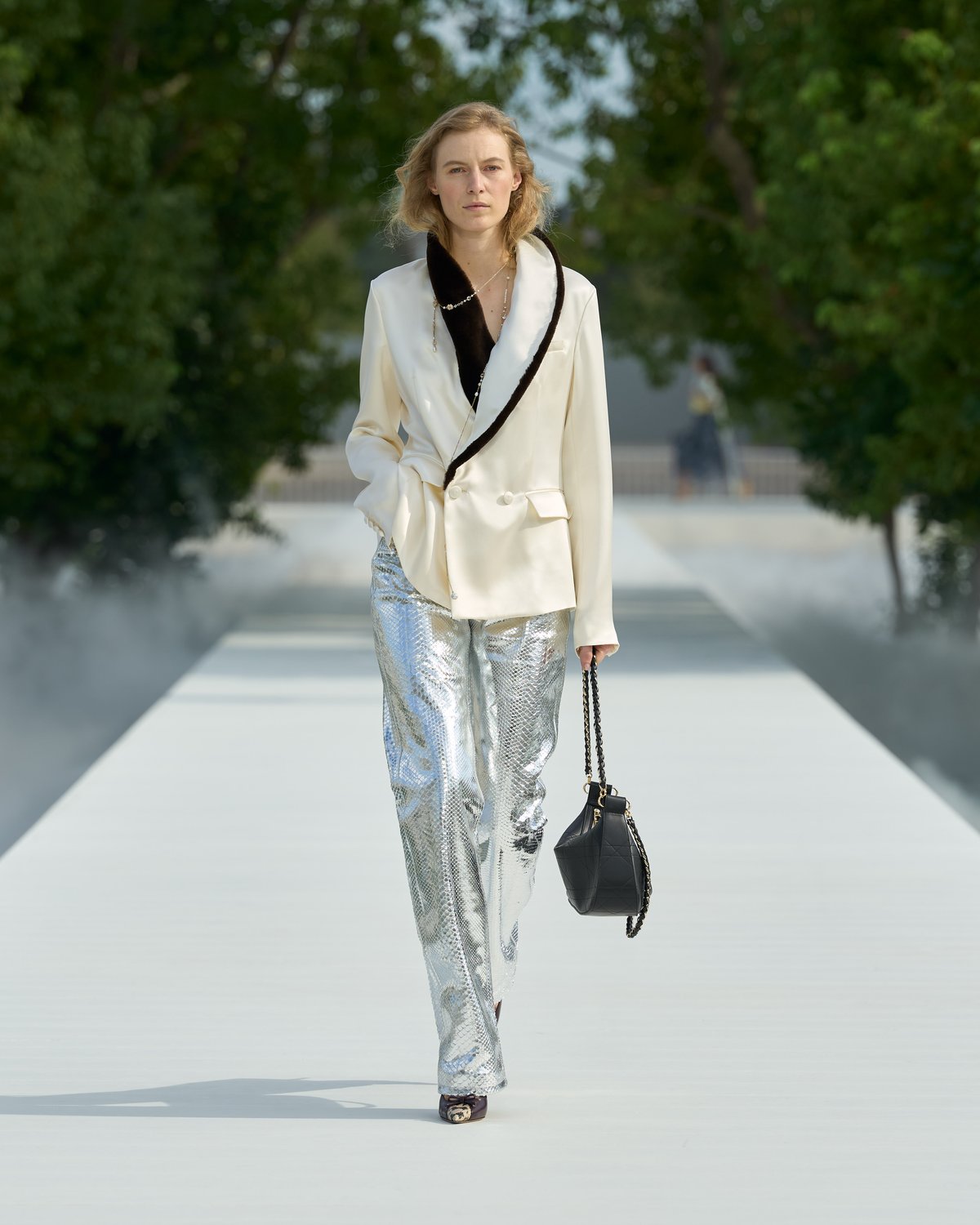 Look 5 from Dior Spring/Summer 2027: a model in an ivory jacket with a black shawl collar and wide metallic silver trousers, carrying a black shoulder bag