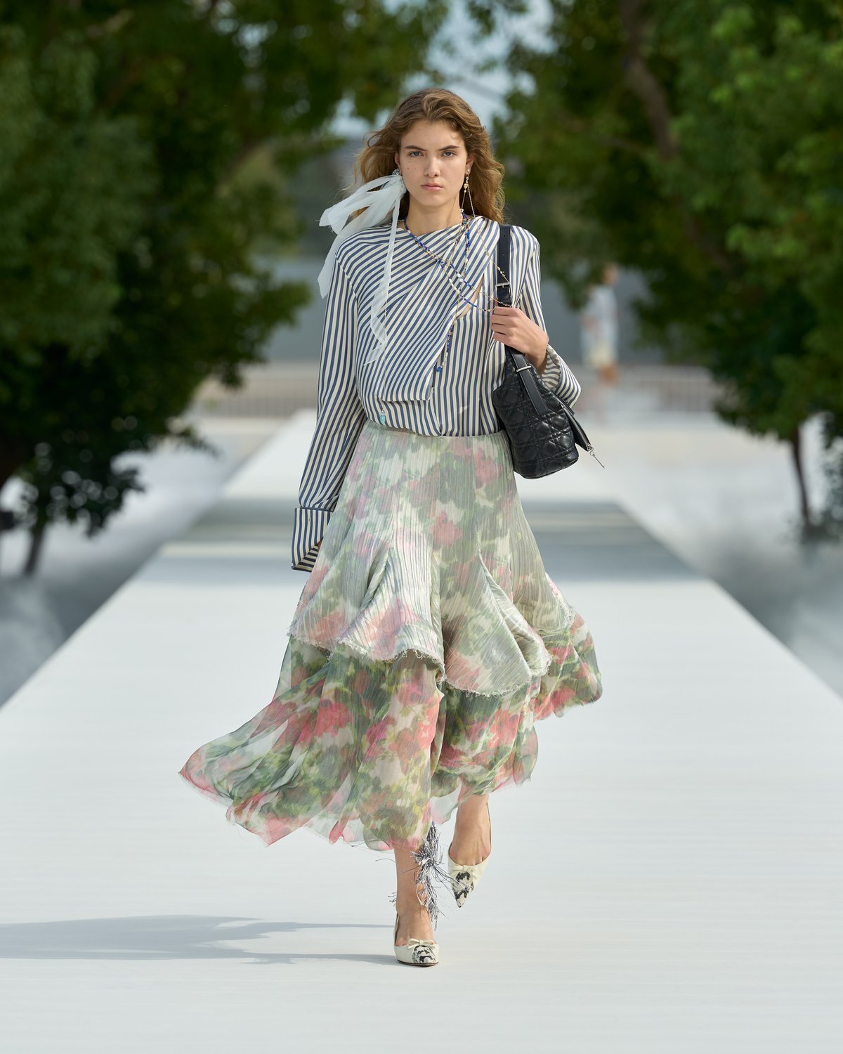 Look 8 from Dior Spring/Summer 2027: a model in a blue and white striped shirt with a white scarf and a full, tiered floral skirt in pink and green, with a black bag on her shoulder