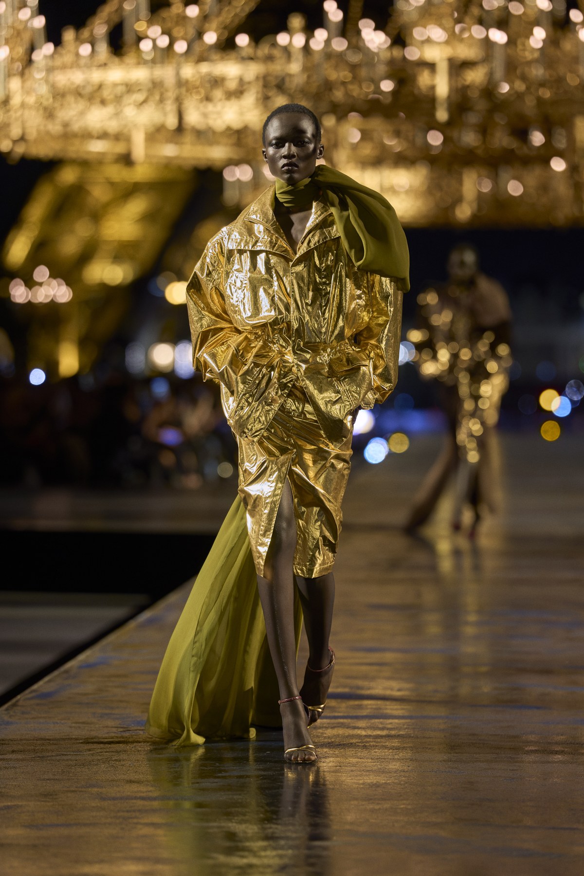 Model in a gold metallic jacket and skirt with an olive-green scarf trailing to the floor on the Saint Laurent Women