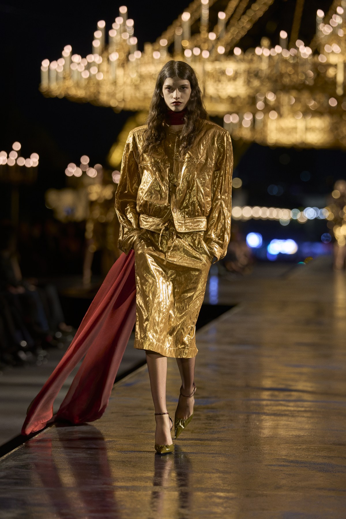 Model in a cropped gold metallic jacket and pencil skirt with a deep red drape falling behind her at the Saint Laurent Women
