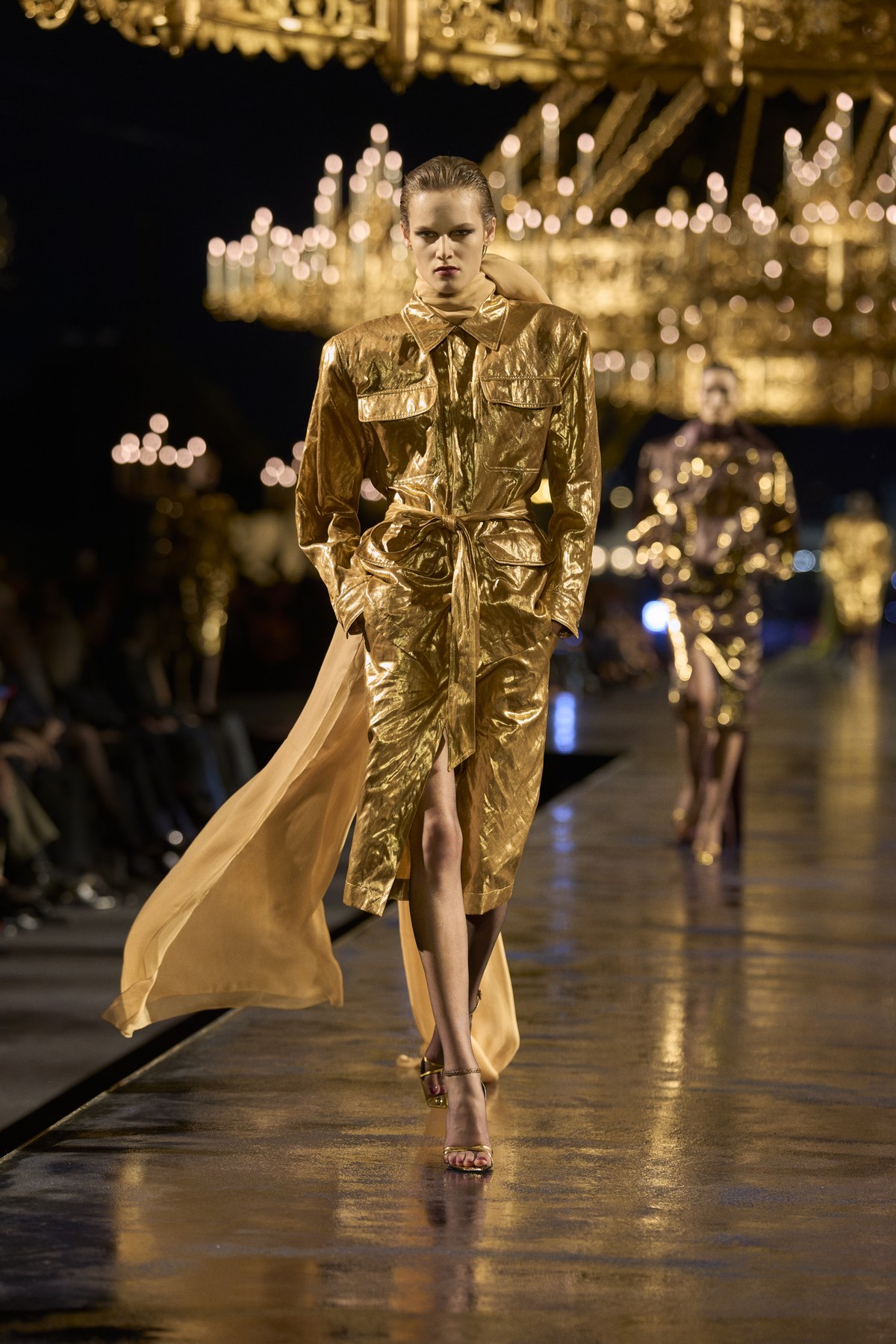 Model in a gold safari-style jacket with patch pockets and a belted skirt, trailed by a long pale gold sash, at Saint Laurent Women