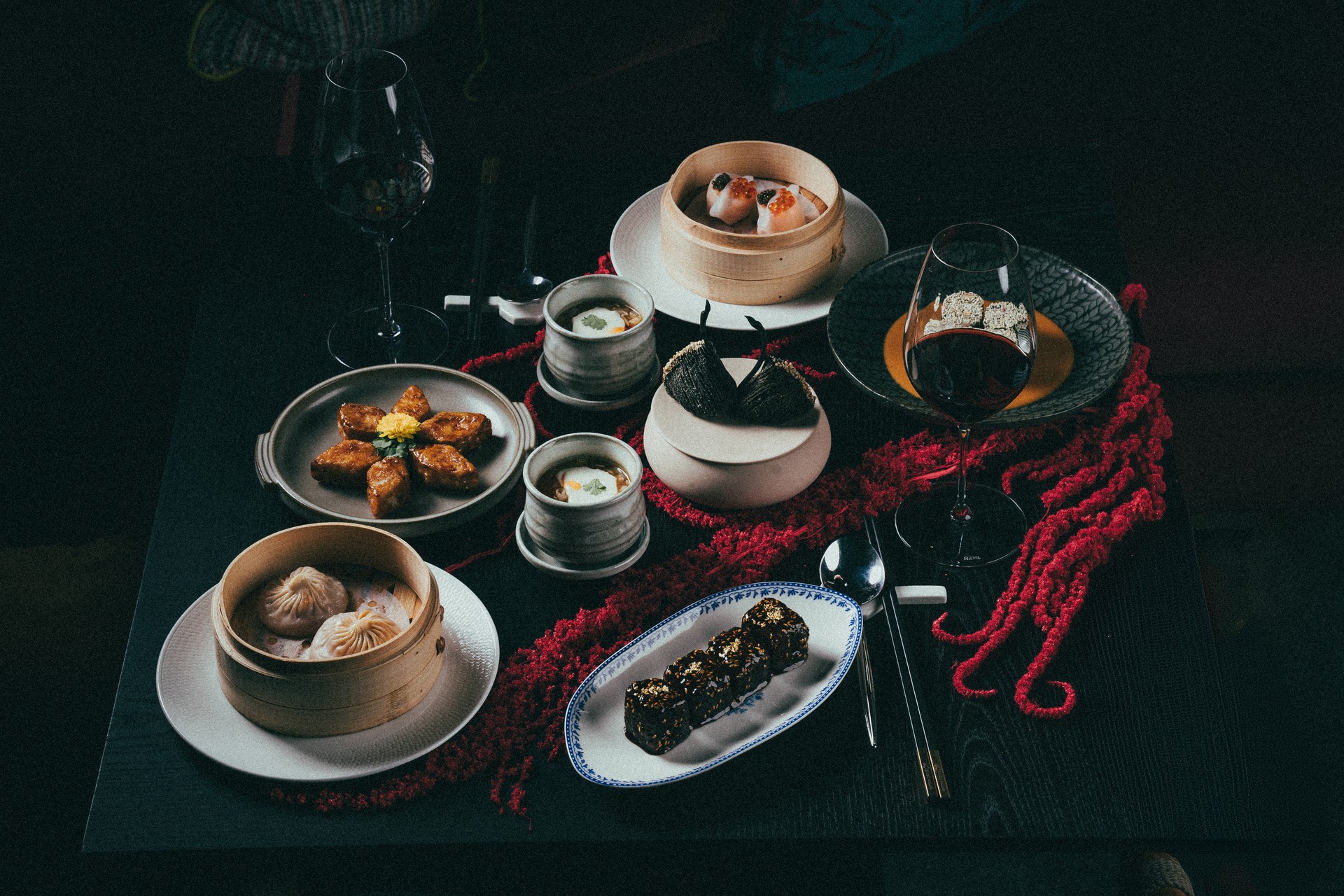 A dark table at Yù & Mì set with bamboo steamers of dumplings, xiao long bao, gold-leaf topped bites, soup cups and glasses of red wine over a red floral garland