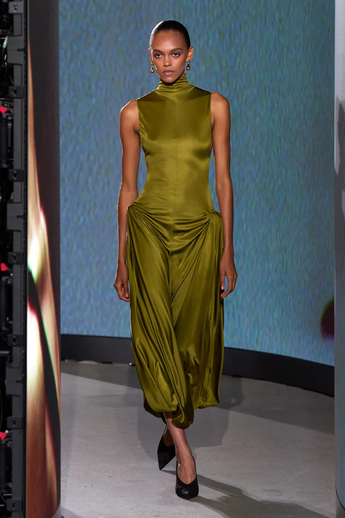 Model on the Alaïa Winter Spring 2027 runway in a high-neck olive satin dress with hip gathers, gold earrings and black pumps between curved LED screens