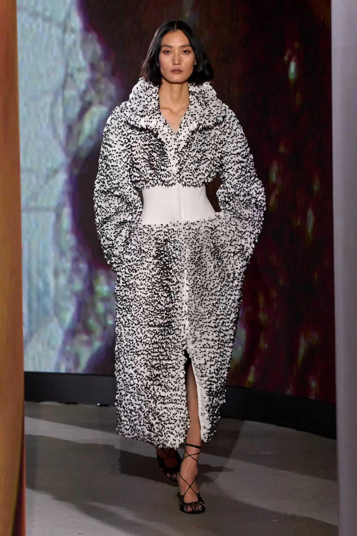 Model on the Alaïa Winter Spring 2027 runway in a white textured jacket and skirt dense with black beads, white knit waistband and black strappy sandals