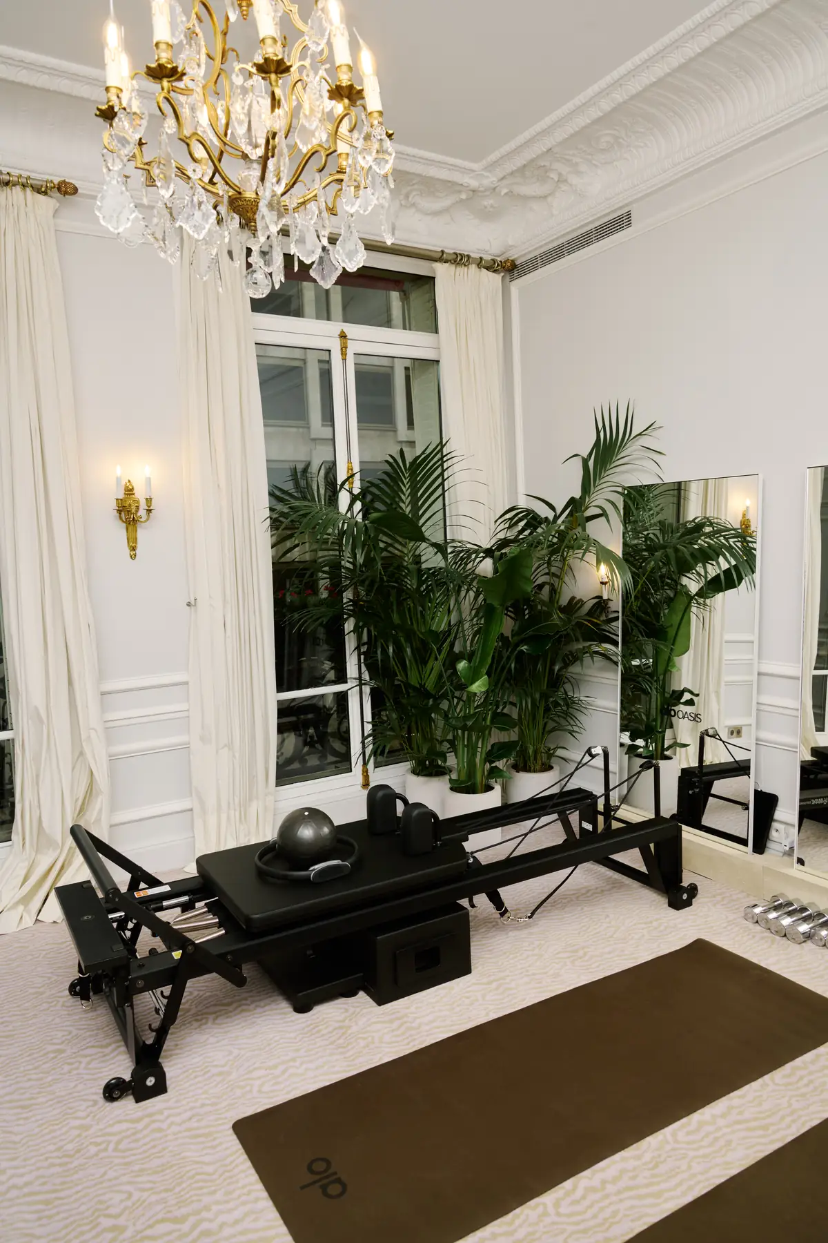 A black Pilates reformer and ALO mat beneath a gilded chandelier in a palm-lined ALO Oasis room during the ALO Plaza Athénée takeover