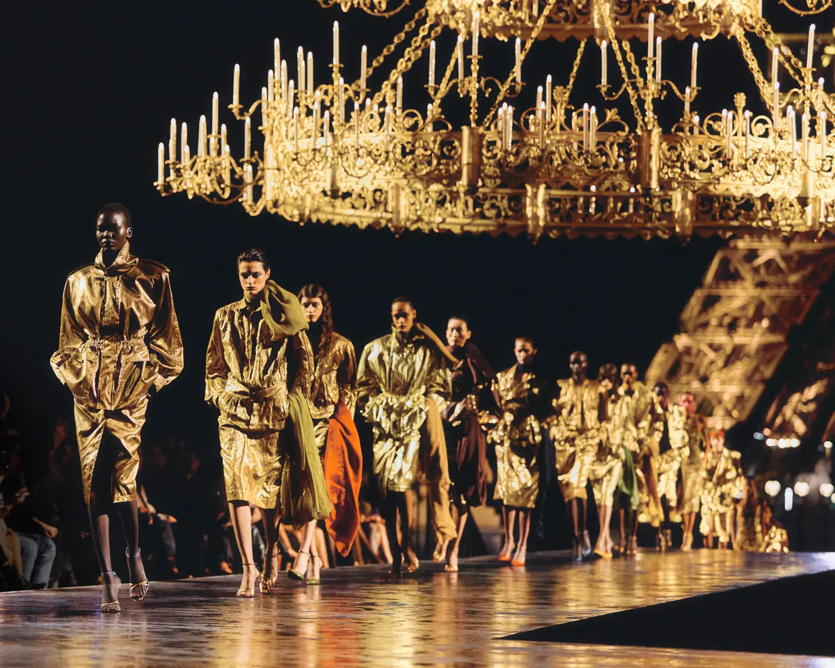 Models in gold looks walk in a line beneath chandeliers at Anthony Vaccarello