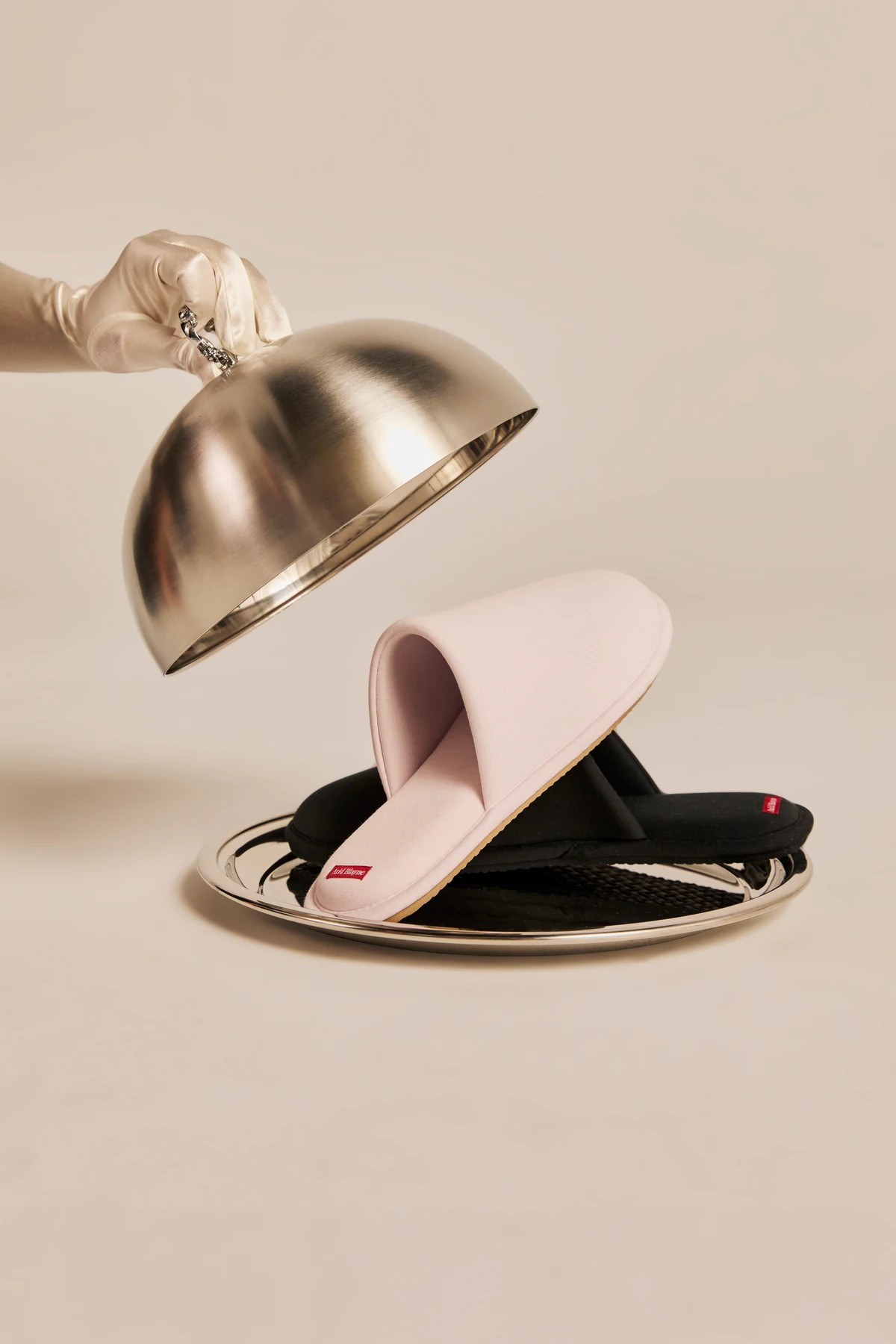 A white-gloved hand lifts a silver cloche to reveal Arid Blayne The Slipper in Blush stacked on a black pair on a silver tray