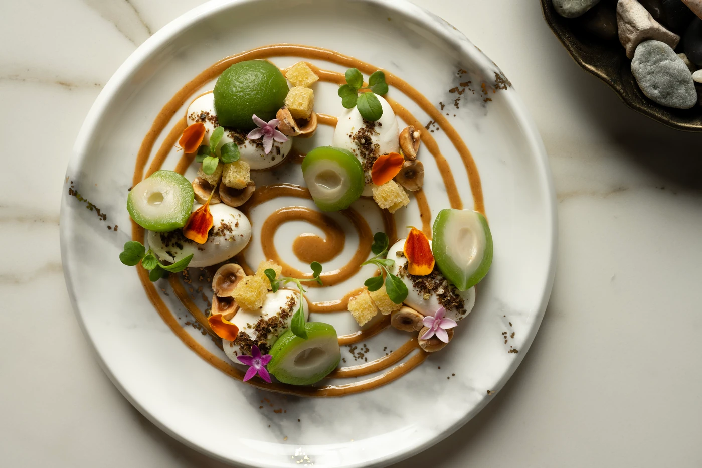 An artfully plated dish with a spiral of sauce, white cream, green fruit, hazelnuts and edible flowers at Bazaar Meat Miami Beach