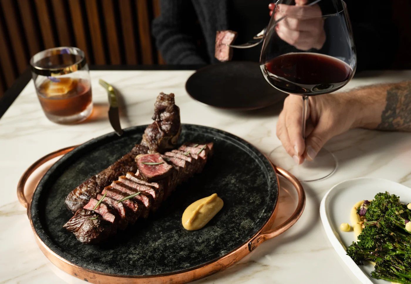 A sliced medium-rare bone-in steak on a stone platter with rosemary and mustard at Bazaar Meat Miami Beach, with red wine and charred broccolini