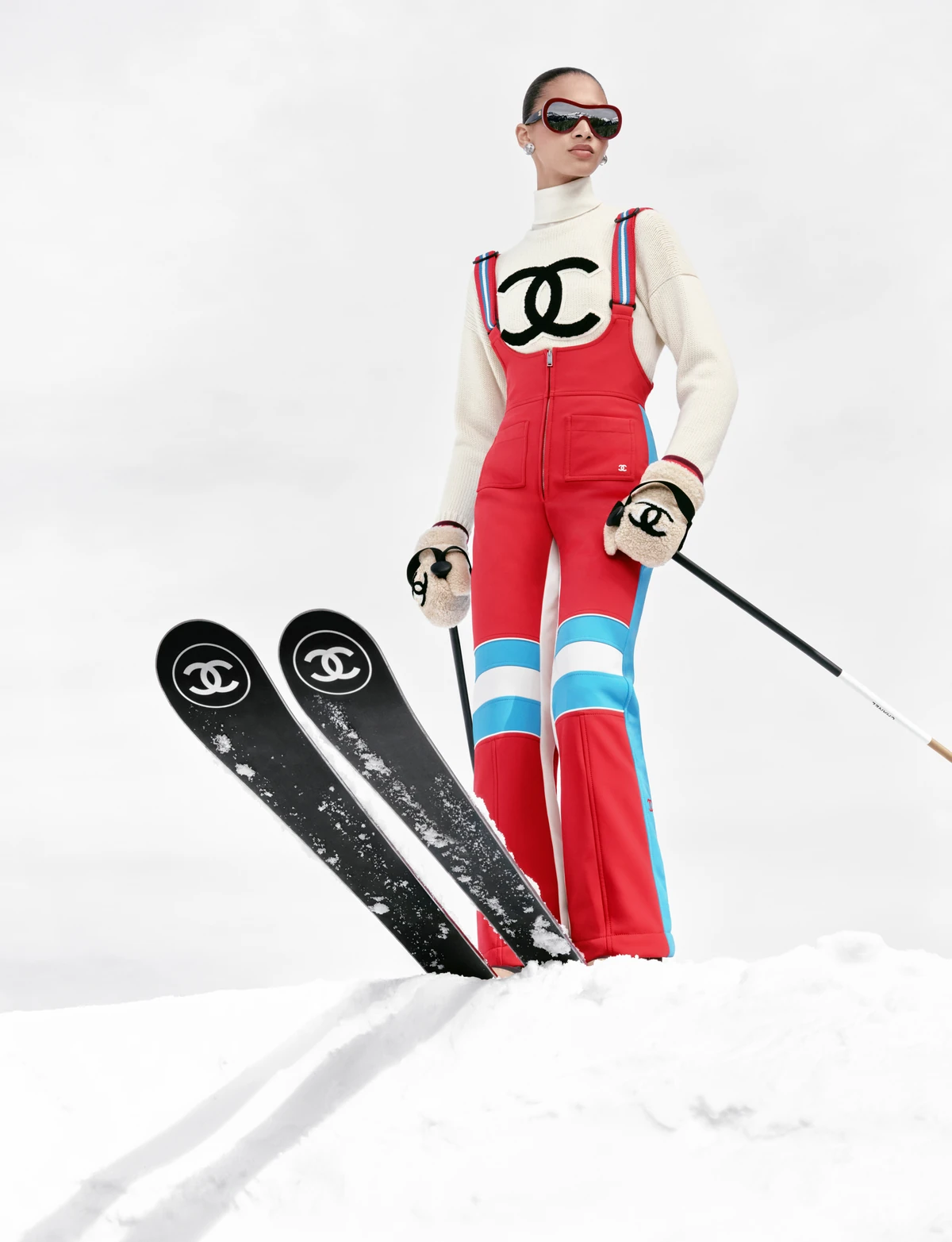 Model in a red Chanel ski suit with blue stripes and a CC logo, holding Chanel skis on a snowy slope, Chanel Coco Neige 2026/27