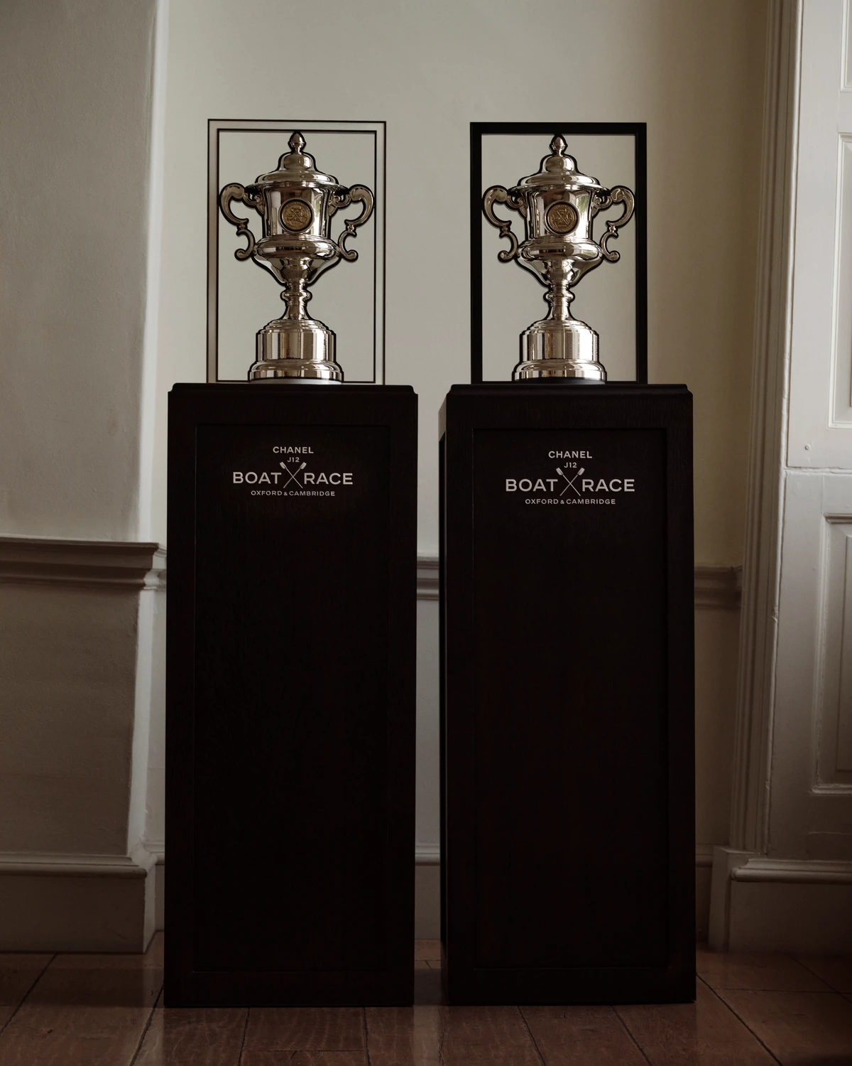 The two silver Chanel J12 Boat Race trophies in white and black rectangular frames on tall black plinths marked Chanel J12 Boat Race Oxford & Cambridge