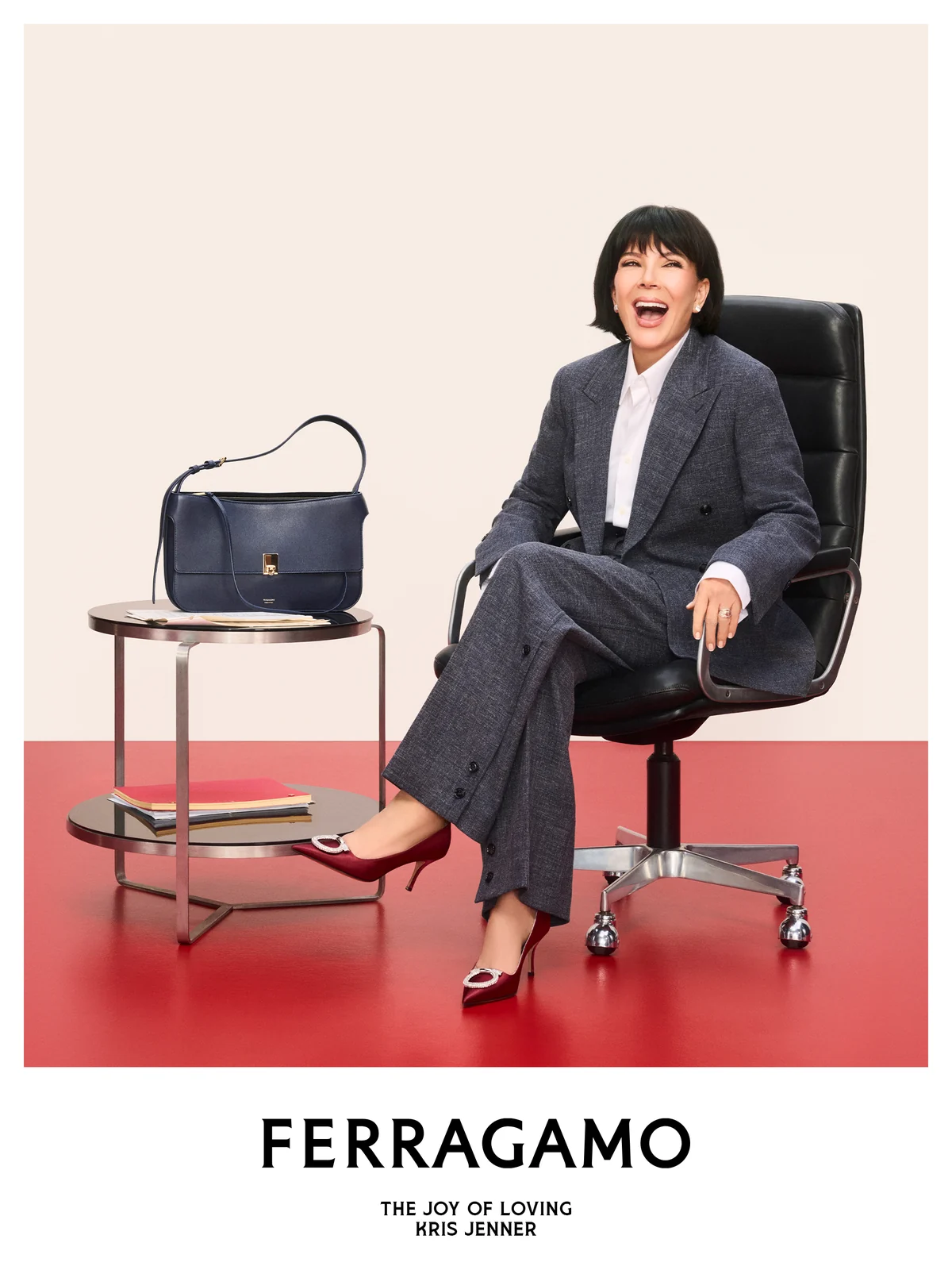 Kris Jenner in a gray suit and red pumps beside a navy Ferragamo bag on a side table in the Ferragamo holiday campaign