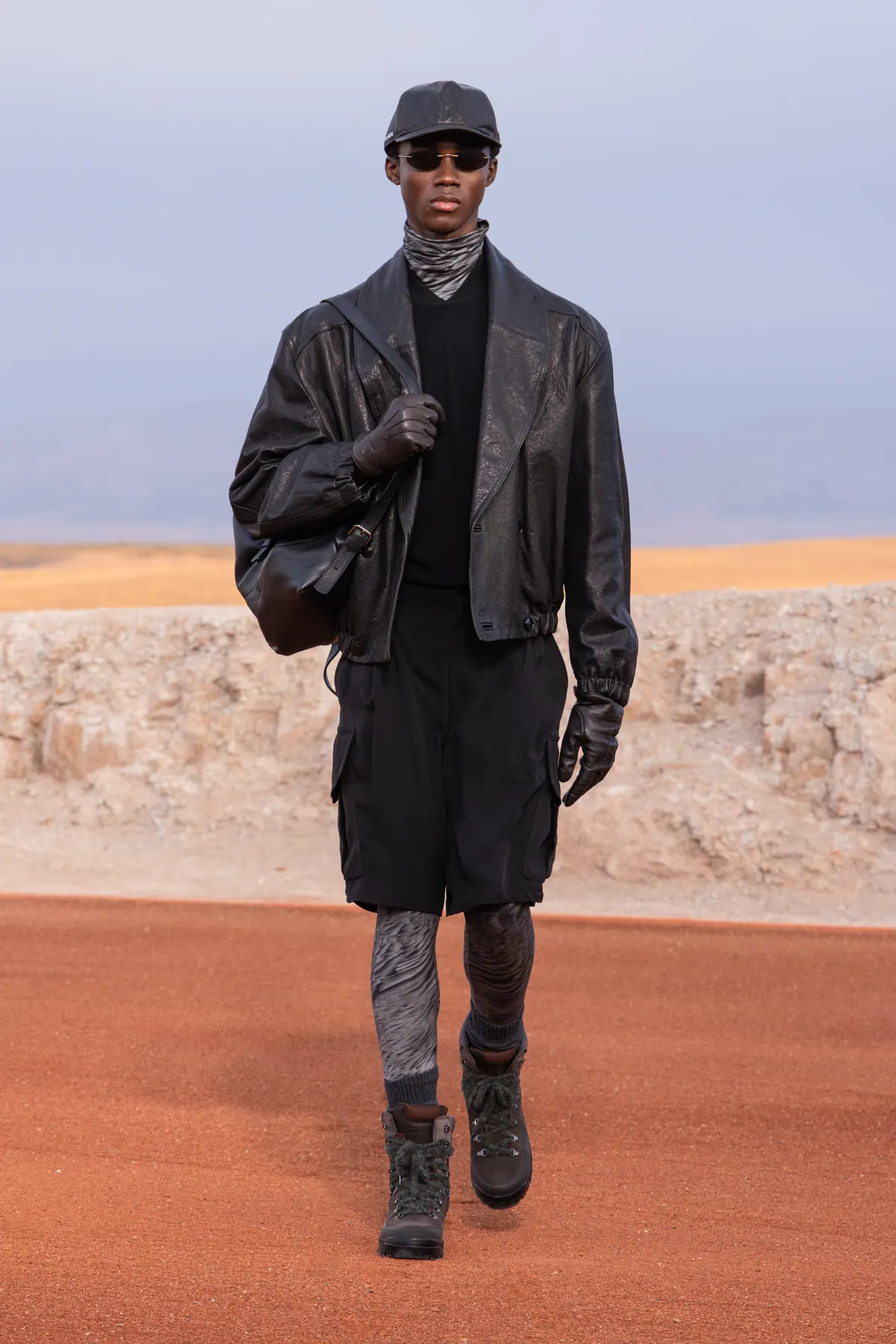 A Giorgio Armani & Kith Dreams of the Dune look: black leather jacket over long black layers with boots, sunglasses and a cap on the desert runway