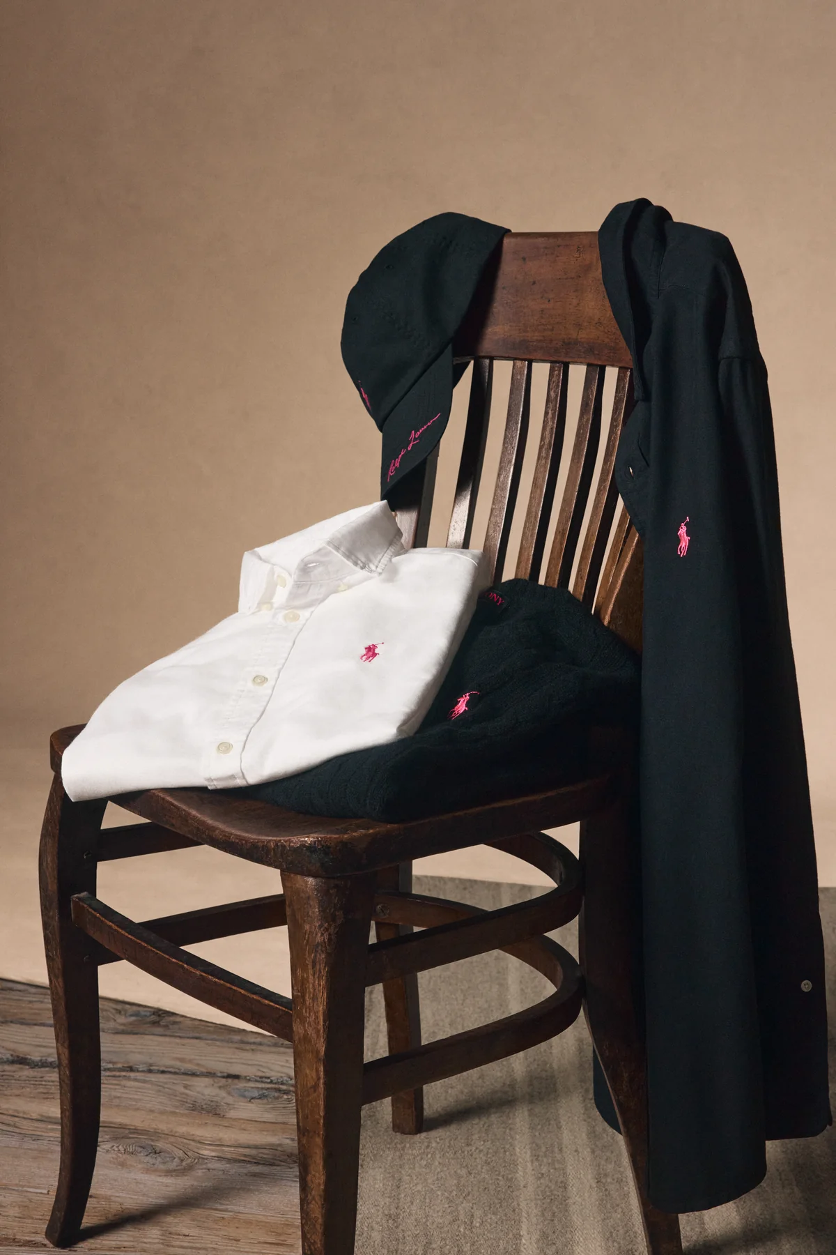 A black Pink Pony Twill Ball Cap, white and black Pink Pony Oxford Shirts and a black cable-knit sweater arranged on a wooden chair, the 100 percent donation pieces in the Ralph Lauren Pink Pony 2026 collection