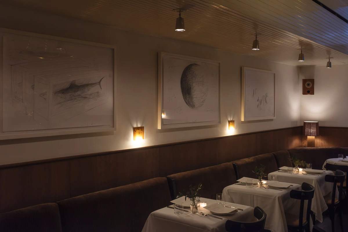 Brown banquette seating and candlelit tables set with white linens beneath framed drawings, including a shark sketch, in Straker’s Soho dining room