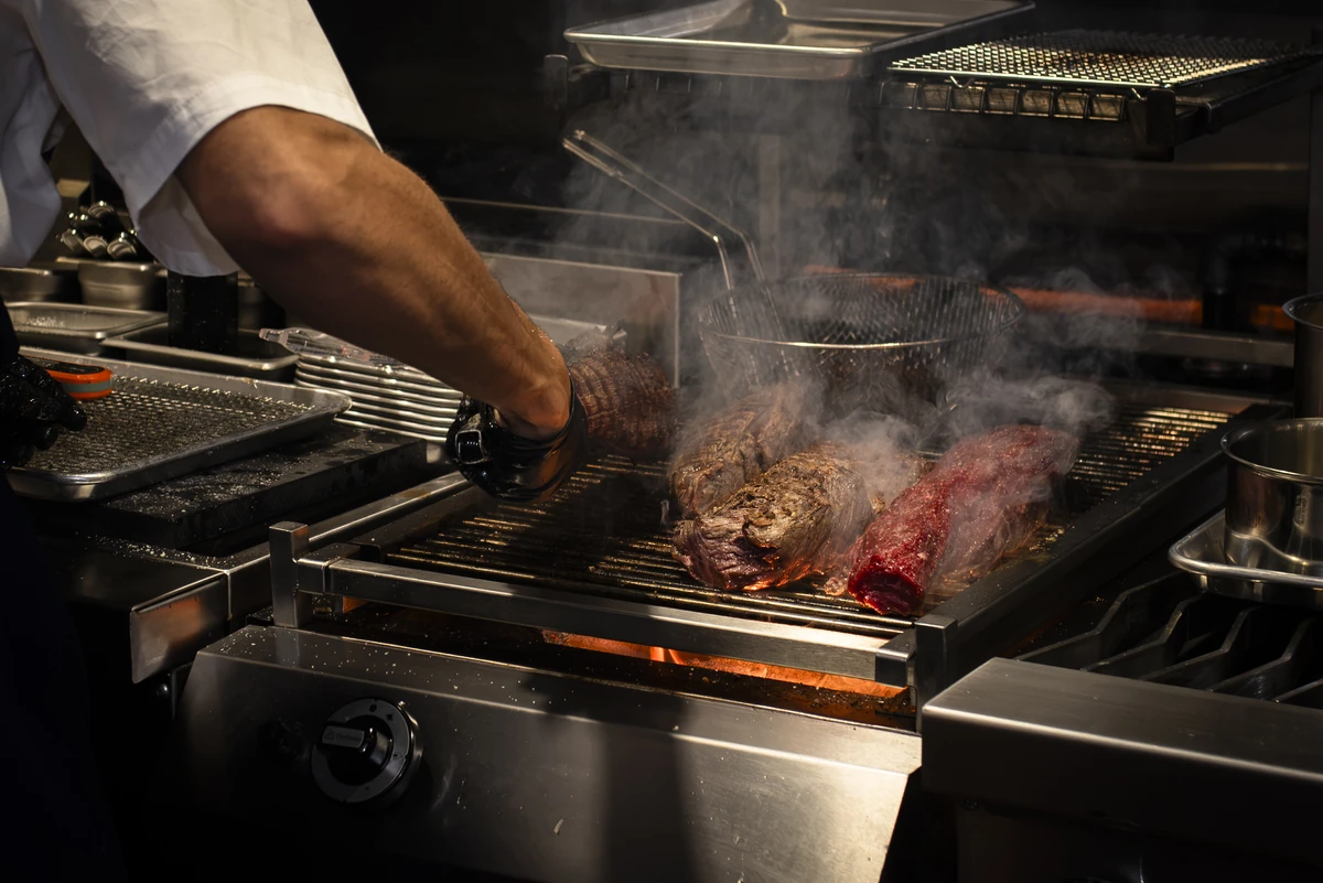A cook’s arm reaching over a smoking open-flame grill with cuts of beef searing in Straker’s open kitchen