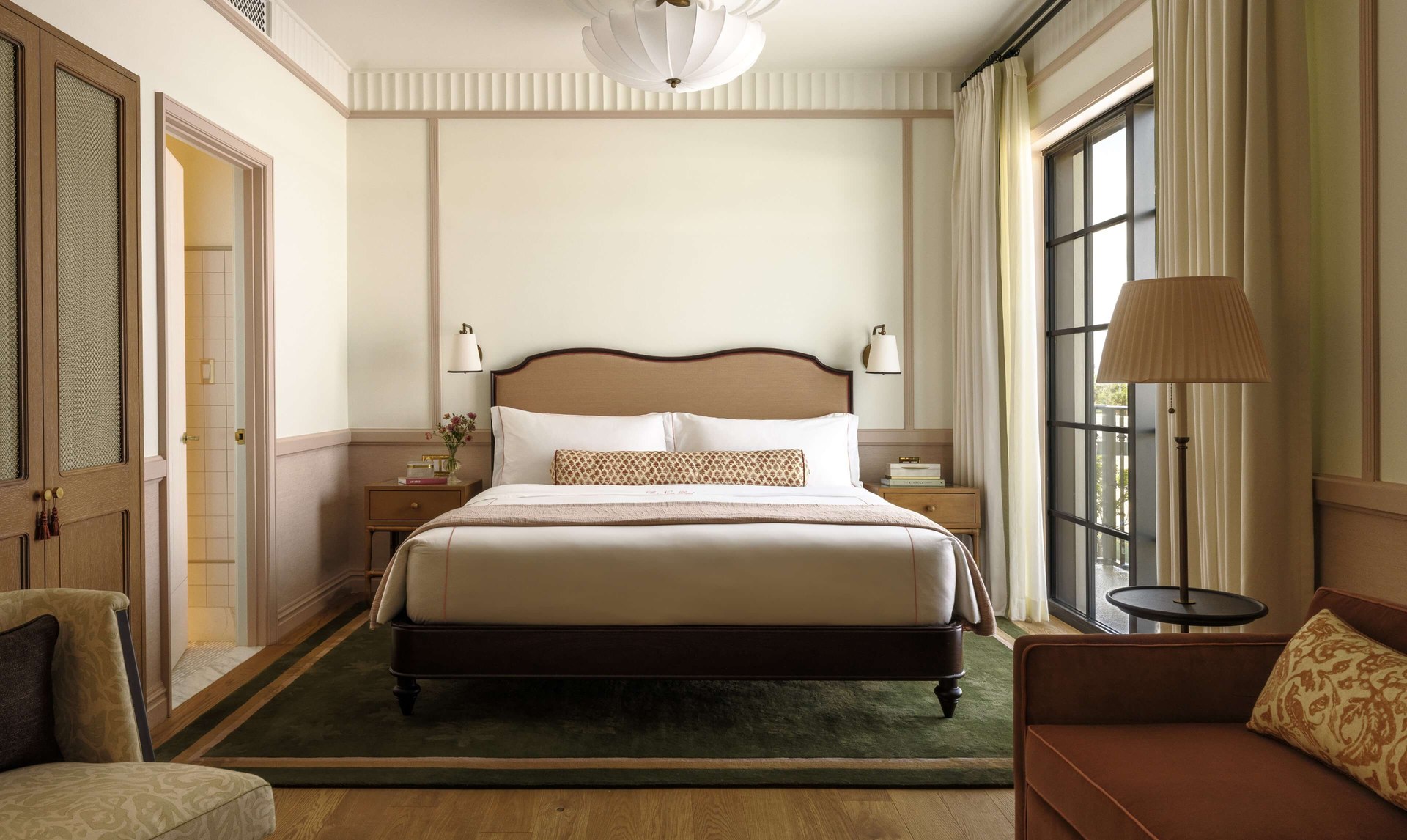 A guest room at The Nora Hotel with an upholstered king bed, green rug, linen-shaded lamp and tall arched windows.